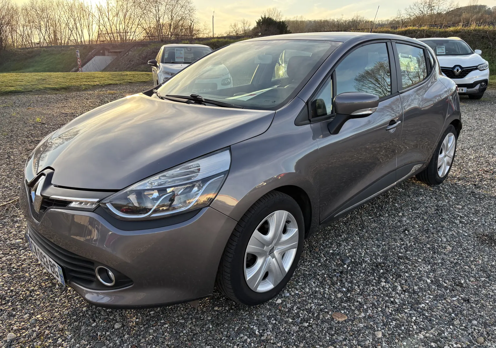 Renault Clio IV gris vue 3/4 avant droit sur parking gravillonné, avec jantes alliage et phares distinctifs.