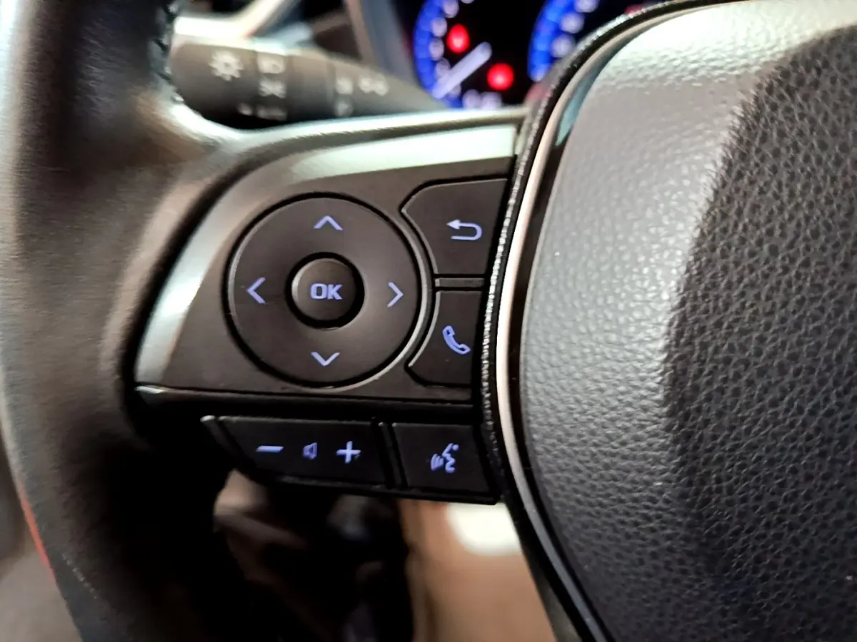 Gros plan sur le volant cuir et alu du Suzuki Swace gris clair, avec commandes multifonctions rétroéclairées en bleu.