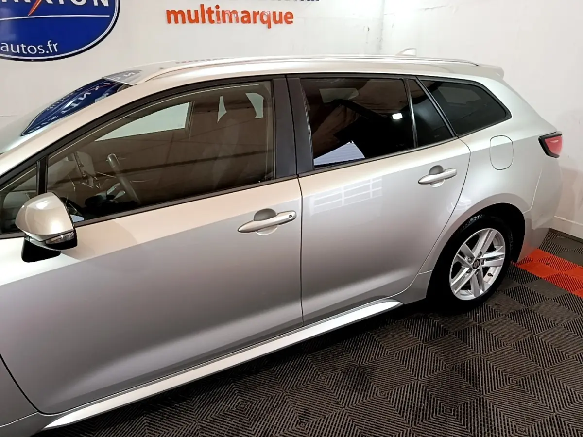 Profil côté gauche de la Suzuki Swace gris clair 2021 avec vitres teintées et jantes alliage visibles en intérieur showroom.