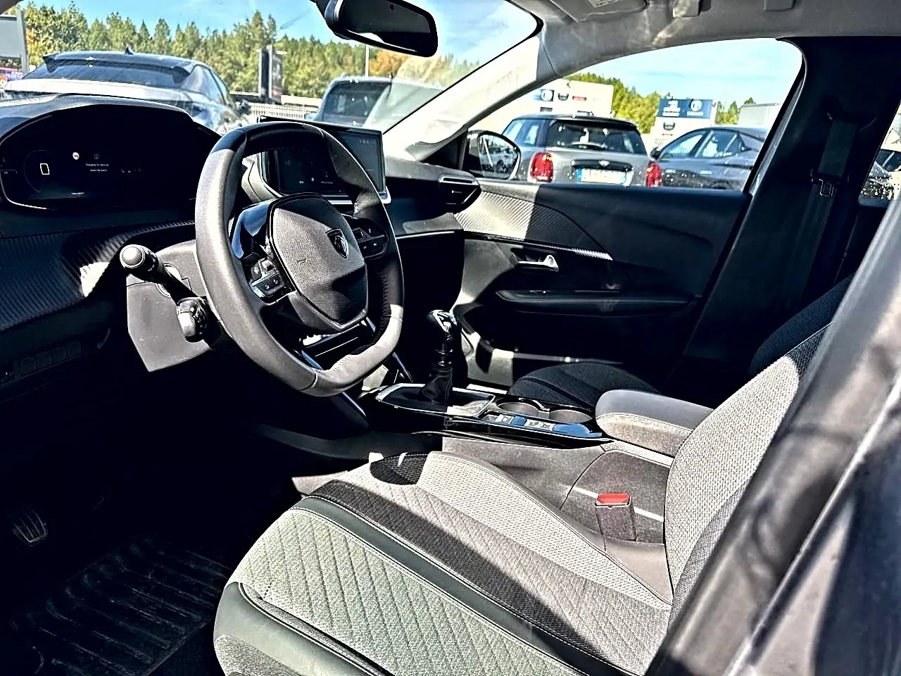 Vue intérieure côté conducteur de la Peugeot 208 1.2 100 Allure 2025 avec volant, tableau de bord et sièges tissu gris.