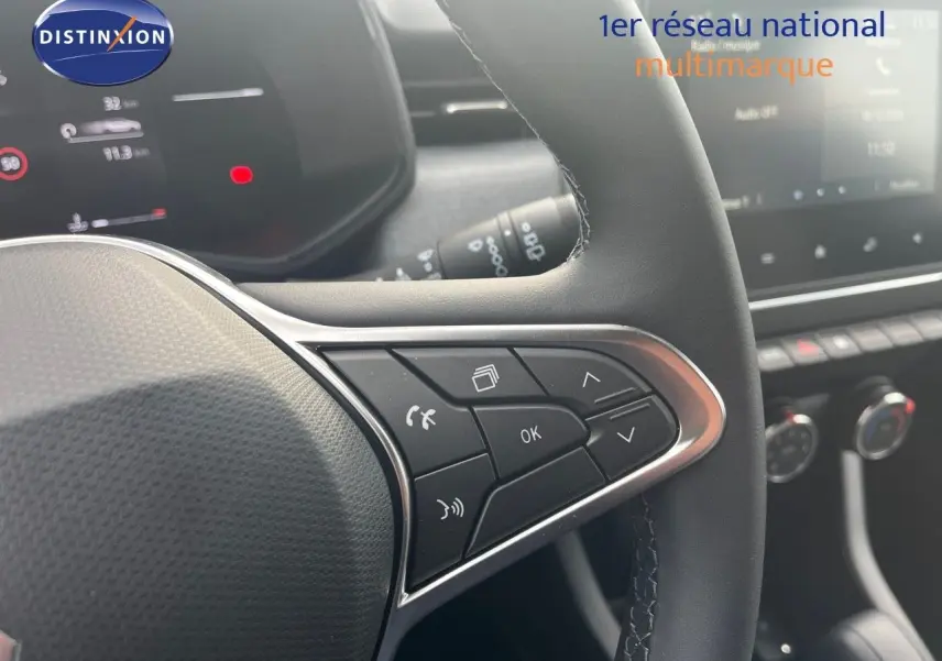 Gros plan sur les commandes au volant côté gauche d'une Renault Clio gris schiste métal, tableau de bord numérique en arrière-plan.