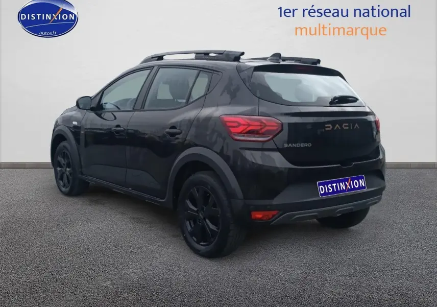 Vue 3/4 arrière droite d'une Dacia Sandero noire avec barres de toit et jantes alu noires, stationnée devant un mur clair.