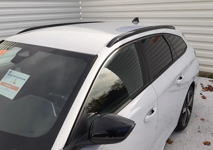 Vue 3/4 avant droit d’une Peugeot 308 SW 2024 blanc okenite avec toit noir et barres de toit noires.
