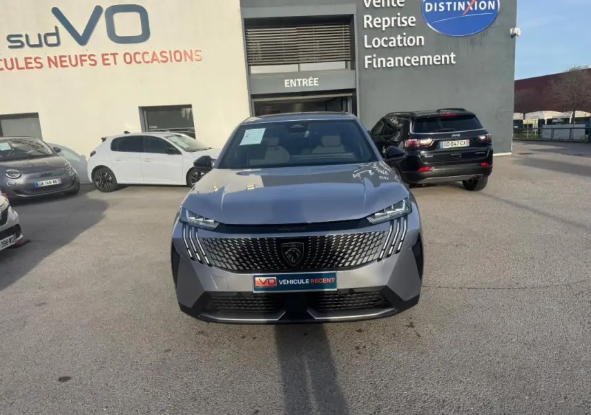 Peugeot 3008 Hybrid gris vue de face, avec calandre distinctive et feux LED allumés, stationné devant un concessionnaire.
