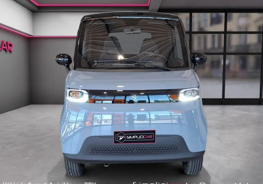 Vue frontale d'une SIMPLICI S1 électrique gris perle avec toit noir et phares allumés dans un showroom moderne.
