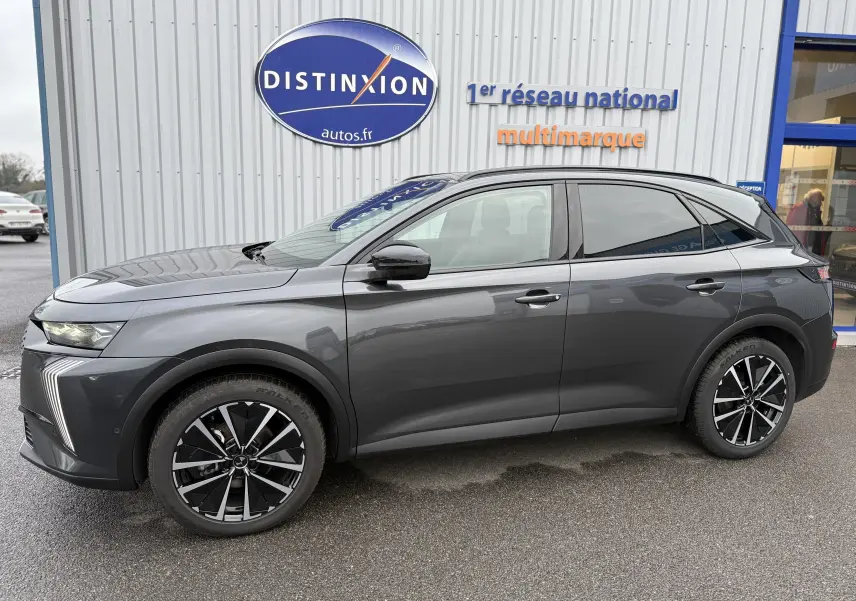 Vue latérale droite du SUV DS7 2025 gris titane avec jantes noires et toit électrique, devant un bâtiment Distinxion.