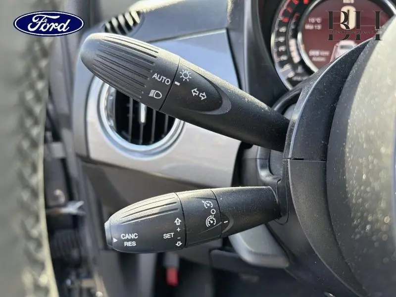 Gros plan sur les commandes au volant noir d'une Fiat 500 Electroclash Grey Métal, avec réglages feux et régulateur de vitesse.
