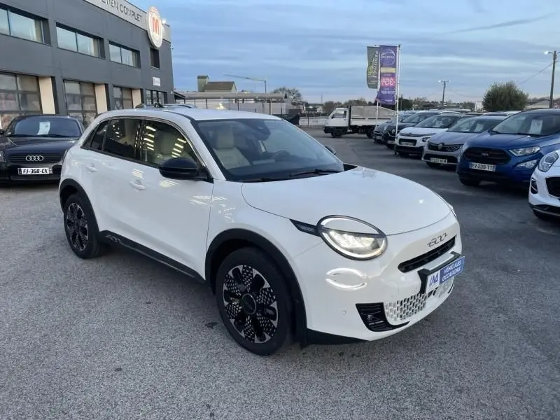 Vue 3/4 avant droite d'une Fiat 600 blanche 2025 avec phares ronds allumés et jantes noires au design moderne.