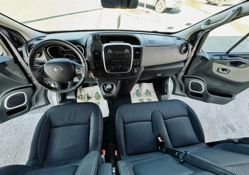 Vue intérieure avant du Nissan NV300 Combi gris, montrant le tableau de bord et les sièges en tissu noir avec portes ouvertes.