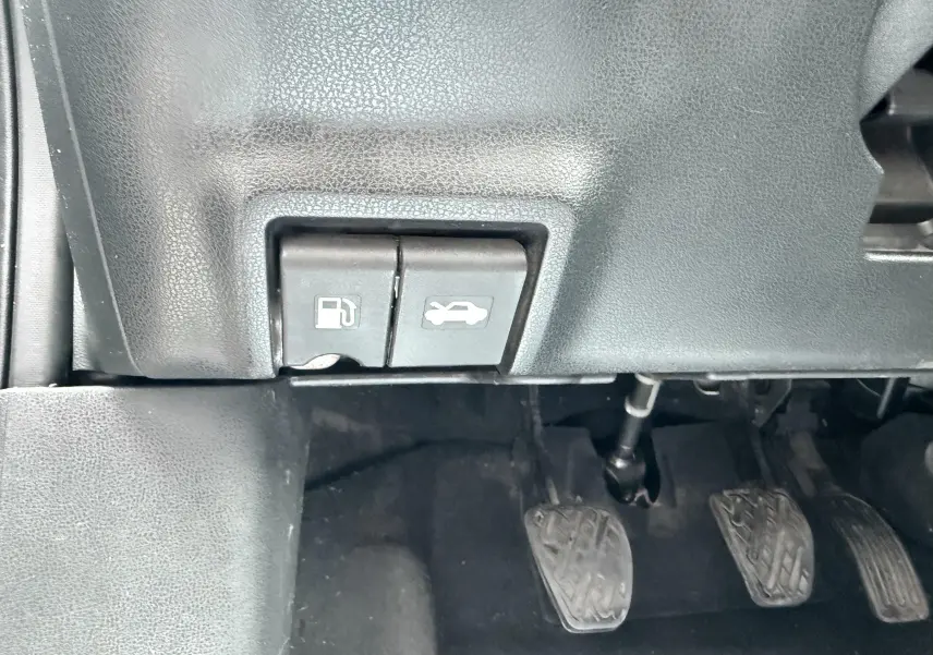 Gros plan sur les commandes d'ouverture de trappe à carburant et capot, avec pédales d'embrayage, frein et accélérateur visibles.