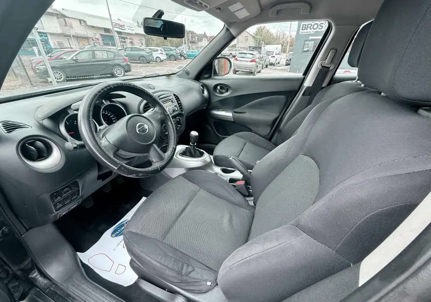 Vue intérieure côté conducteur du Nissan Juke noir 2012, montrant le volant cuir, la console centrale et les sièges tissu noirs.