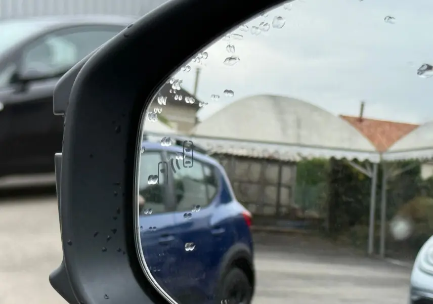 Reflet dans le rétroviseur montrant un SUV bleu en stationnement, avec gouttes de pluie sur le miroir.