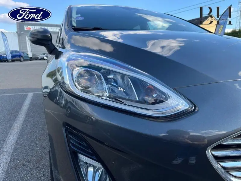 Gros plan sur le phare avant droit d'une Ford Fiesta 2021 gris Fashion Magnetic, avec reflet du ciel sur la carrosserie.