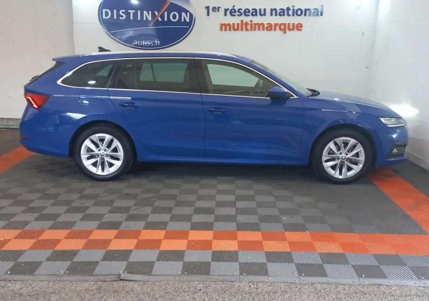 Vue latérale droite d'une Skoda Octavia break bleu 2022 dans un showroom avec sol à damiers gris et orange.