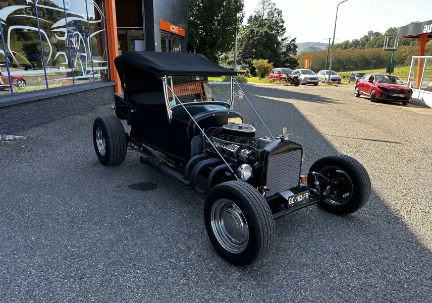 Ford T Hot Rod V8 noir vue 3/4 avant droit, capote relevée et moteur V8 apparent sur parking extérieur.