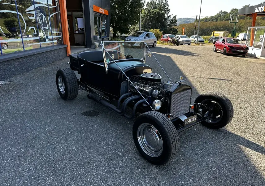Ford T Hot Rod V8 noir en 3/4 avant droit, capote décapotée et moteur V8 apparent sur parking extérieur.