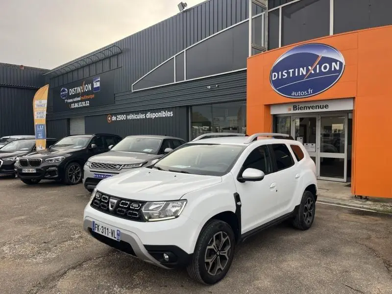 Vue 3/4 avant d'un Dacia Duster blanc glacier 2019 avec jantes alliage et barres de toit chromées devant un concessionnaire.
