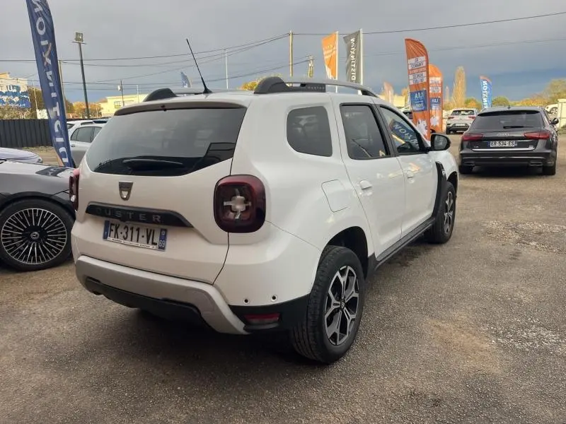 Vue 3/4 arrière droite d'un Dacia Duster blanc Glacier avec jantes alliage et barres de toit chrome satiné.