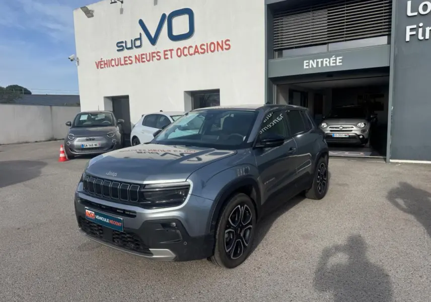 Jeep Avenger gris en 3/4 avant droit, avec jantes alliage noires et calandre distinctive, stationné devant un garage.
