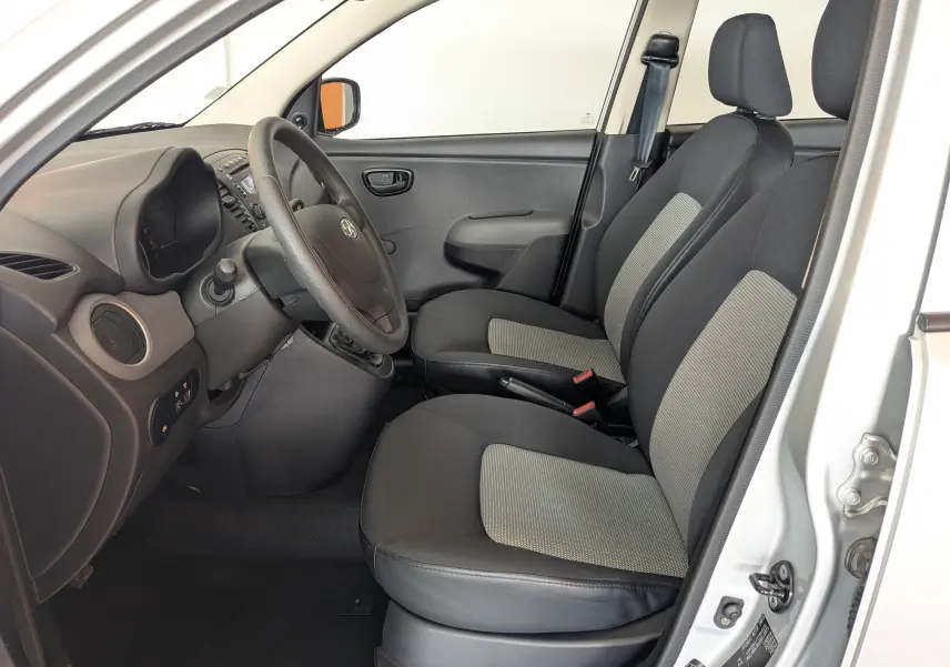 Vue intérieure côté conducteur de la Hyundai i10 gris clair 2010, montrant les sièges tissu gris et tableau de bord noir.