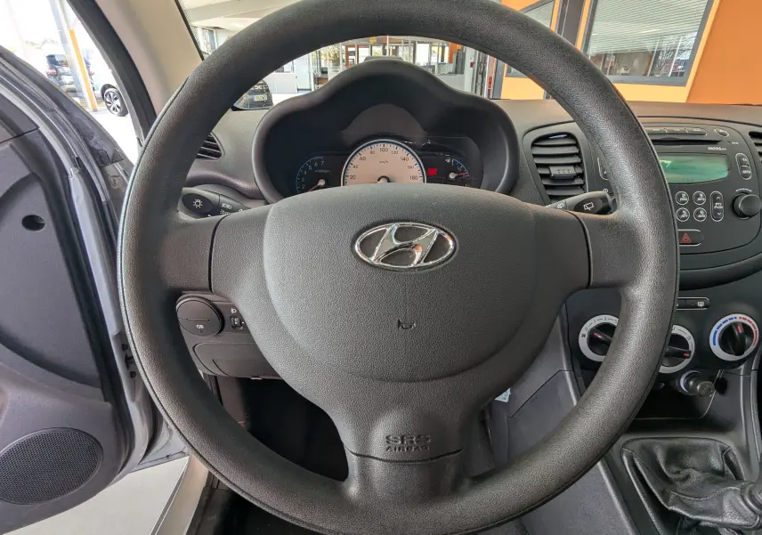 Vue intérieure centrée sur le volant et le tableau de bord d'une Hyundai i10 gris clair de 2010 avec commandes manuelles visibles.