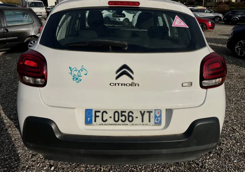 Vue arrière d'une Citroën C3 blanche de 2019 avec un autocollant floral bleu à gauche du logo et un triangle rose sur la vitre.