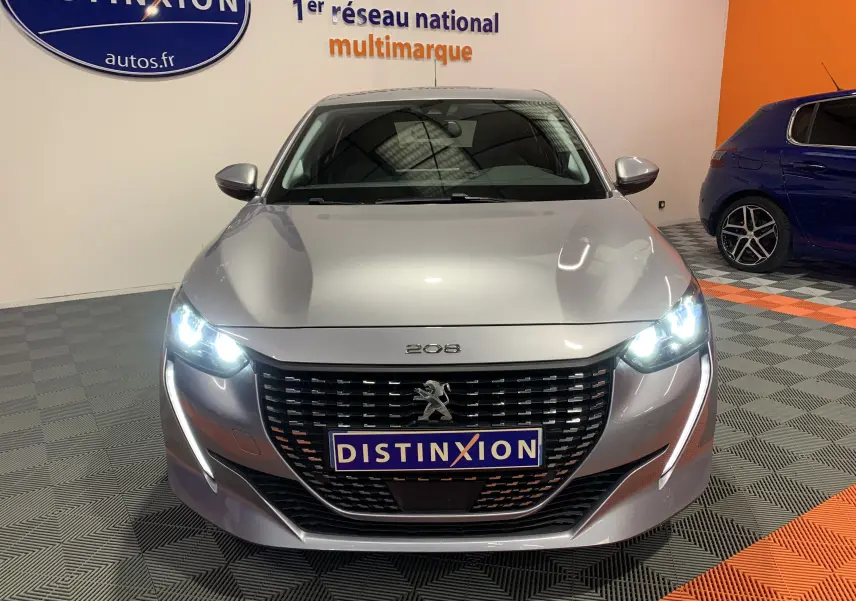 Vue frontale d'une Peugeot 208 gris métallisé avec phares allumés dans un showroom multimarque.