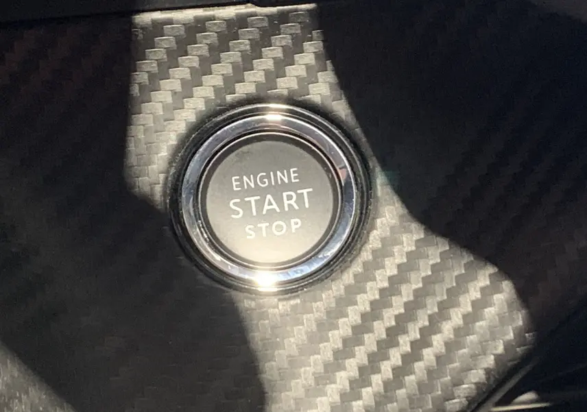 Gros plan sur le bouton start/stop moteur entouré d’un habillage effet carbone dans une Peugeot 208 gris, intérieur visible.