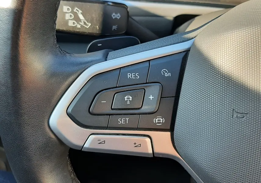 Gros plan sur les commandes au volant côté gauche du Volkswagen T-Roc bleu 2022, avec boutons de régulateur de vitesse et aides à la conduite.