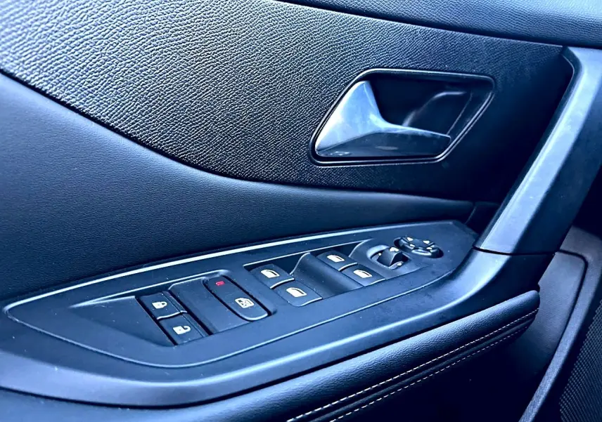 Gros plan sur la commande des vitres et la poignée intérieure côté gauche d’une Peugeot 308 noire, finition 2025.