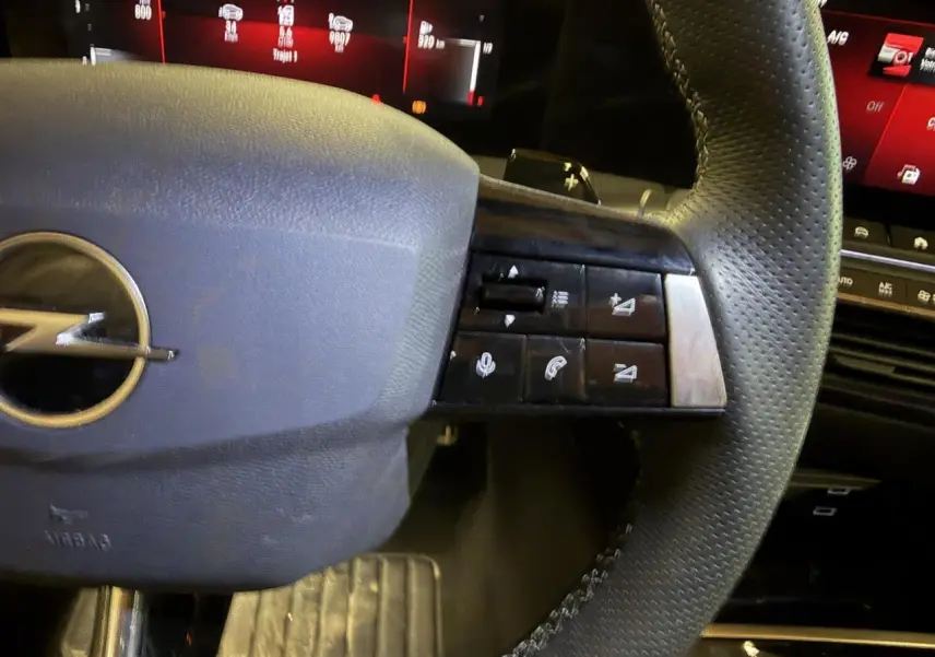 Gros plan sur le volant en cuir perforé de l’Opel Astra 2024, avec commandes multifonctions et tableau de bord digital rouge en arrière-plan.