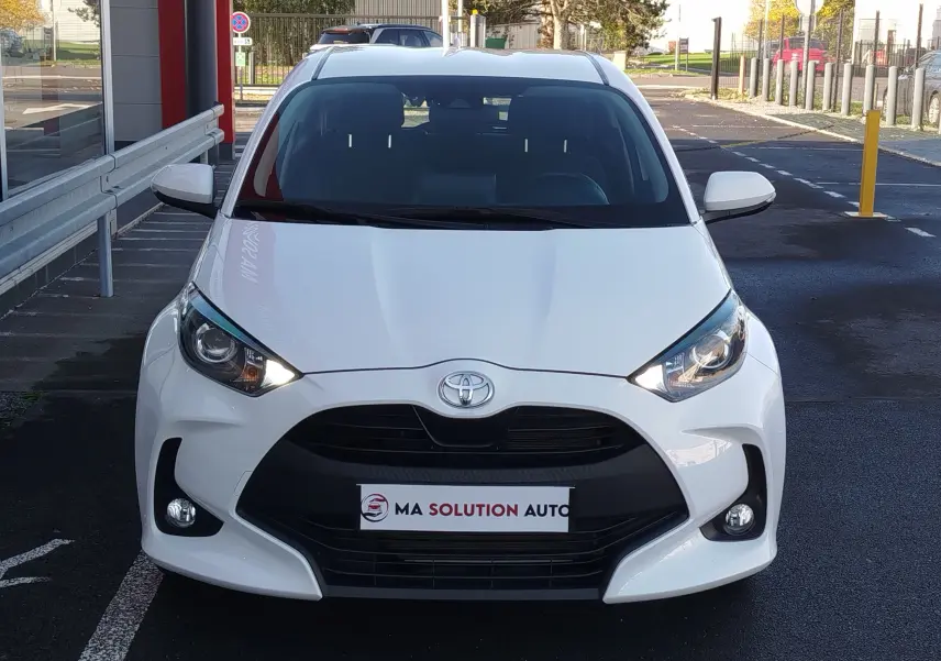 Vue avant d'une Toyota Yaris blanche pur 2023, avec calandre noire et phares allumés, stationnée devant un bâtiment.