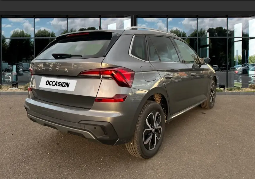 Vue 3/4 arrière droite d'un Skoda Kamiq gris clair avec jantes noires et toit ouvrant, stationné devant une vitrine.