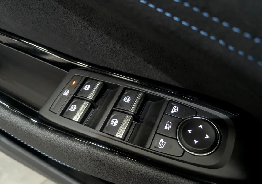 Gros plan sur les commandes des vitres électriques et réglages des rétroviseurs côté conducteur avec surpiqûres bleues sur la garniture.
