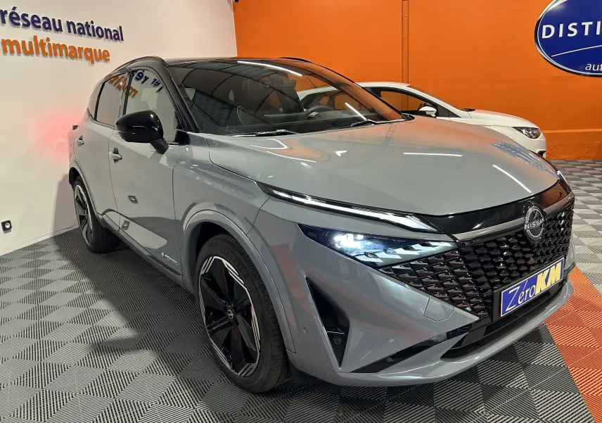Nissan Qashqai gris céramique métal vu en 3/4 avant droit avec phares allumés et calandre noire distinctive.