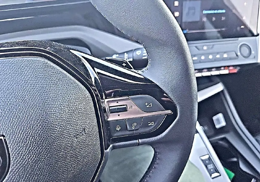 Gros plan sur le volant multifonction noir de la Peugeot 308 SW 1.5 BlueHDi 130 Allure gris Artense, avec commandes audio visibles.