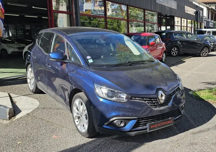 Renault Scenic bleu cosmos métal vu en 3/4 avant droit devant un garage AD Expert avec drapeau promotionnel.