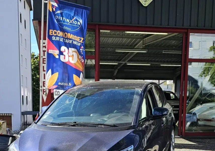 Renault Scénic bleu cosmos métal en 3/4 avant droit, garé devant un garage avec plaque SAGE AUTOMOBILE visible.