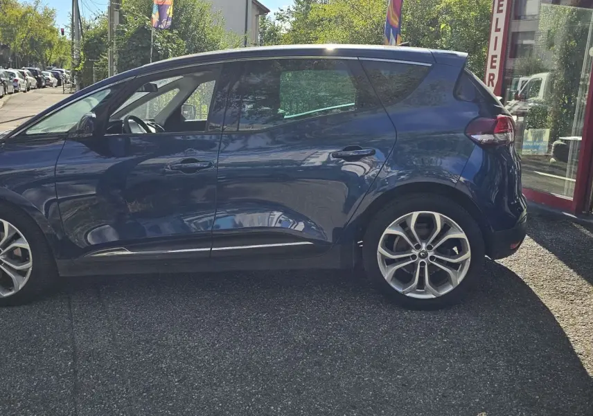 Profil côté gauche d'un Renault Scénic bleu cosmos métal 2020 garé devant un atelier avec jantes alliage visibles.