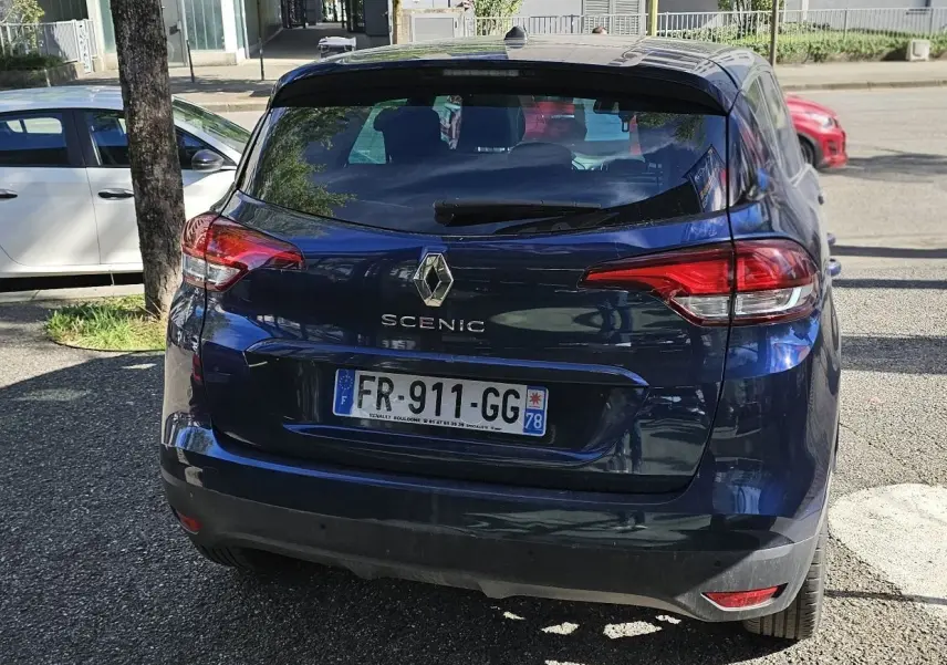 Vue arrière d'un Renault Scénic bleu cosmos métal garé, avec feux arrière rouges et plaque d'immatriculation française.