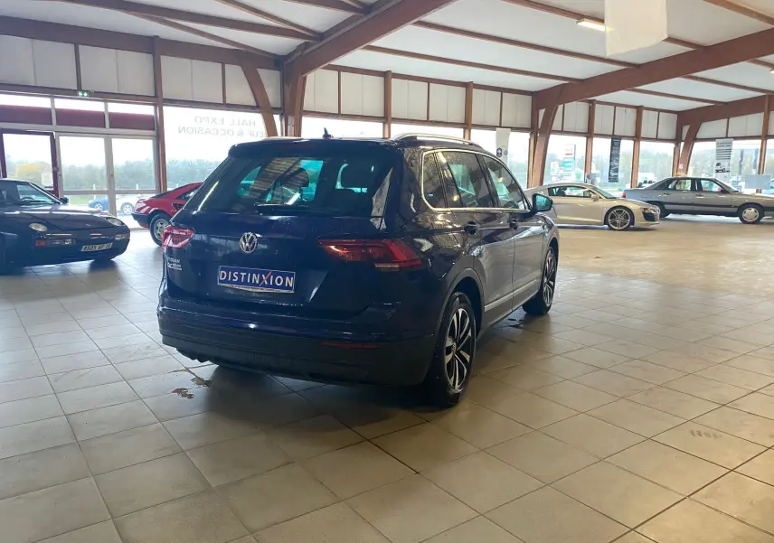 Vue 3/4 arrière d’un Volkswagen Tiguan bleu vert 2020 dans un showroom lumineux avec plusieurs autres voitures visibles.