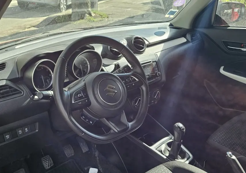 Intérieur de la Suzuki Swift 1.2 Dualjet Hybrid 2023 en rouge, vue côté conducteur avec volant et tableau de bord visibles.