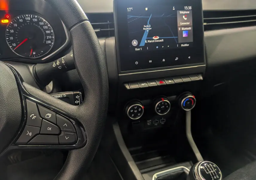 Vue intérieure centrée sur le volant et la console centrale avec écran tactile et levier de vitesses manuel d'une Renault Clio V blanche.
