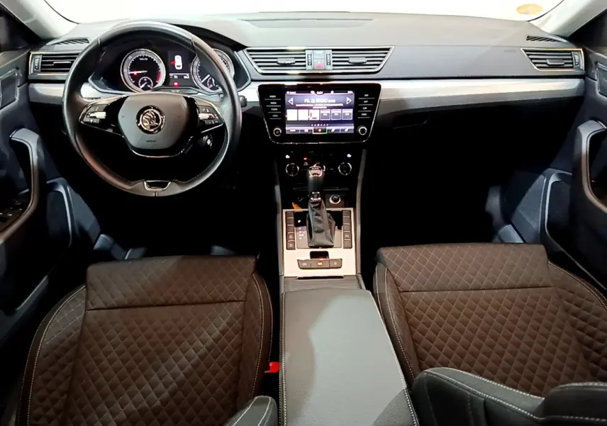 Intérieur de la Skoda Superb 2020 vu de face, tableau de bord noir avec écran tactile et sièges en tissu matelassé.