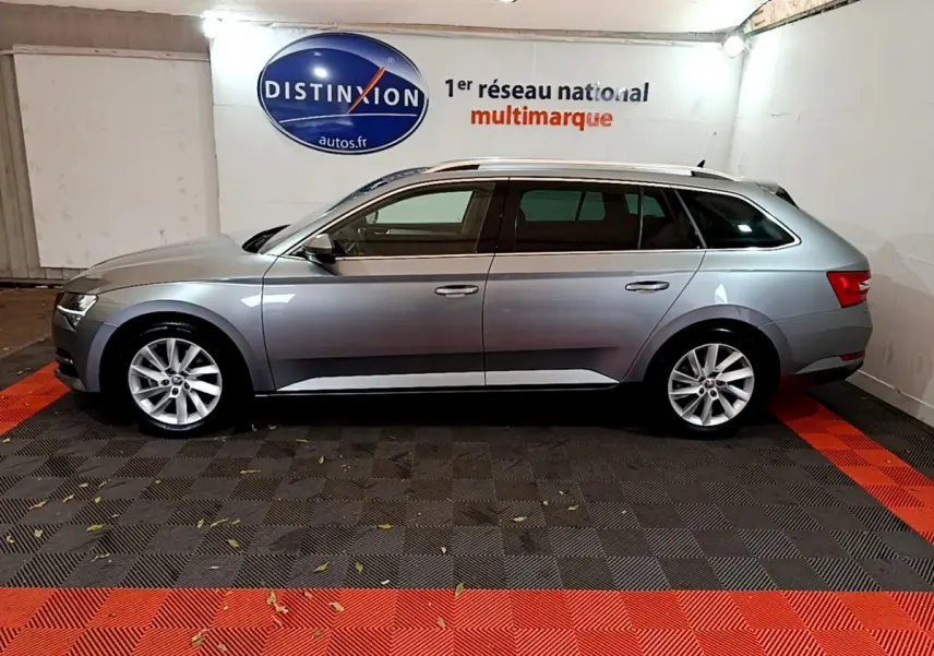 Profil latéral droit d'une Skoda Superb gris clair en version break, stationnée en intérieur sur sol noir et rouge.
