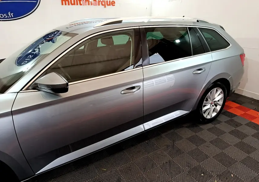 Profil côté gauche d'une Skoda Superb gris clair 2020 en stationnement intérieur sur sol damier noir et rouge.