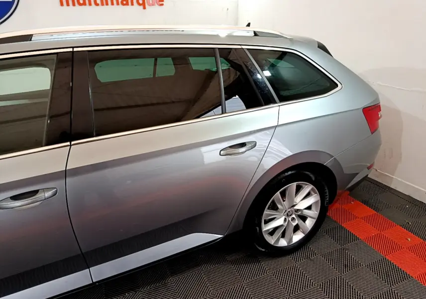 Profil côté gauche d'une Skoda Superb gris clair stationnée sur un sol à motifs noirs et rouges en intérieur.