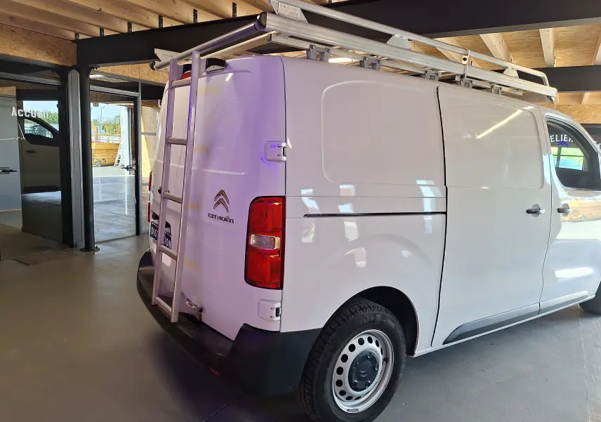 Vue 3/4 arrière droite du Citroën Jumpy blanc avec galerie et échelle métallique sur le toit, dans un garage.