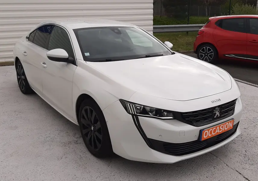 Peugeot 508 blanc vue 3/4 avant droit, calandre noire distinctive et jantes foncées, stationnée devant un mur blanc.