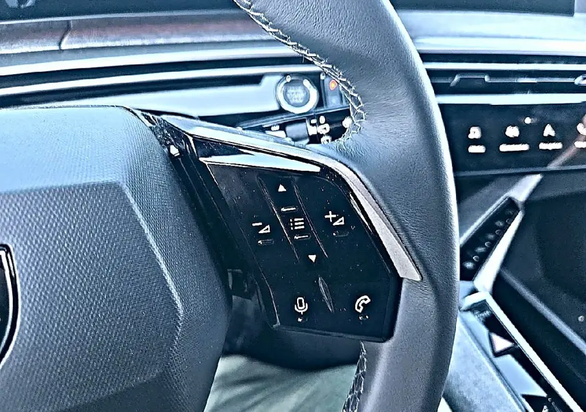 Gros plan sur le volant cuir noir multifonction du Peugeot 5008 blanc, avec commandes vocales et audio visibles.